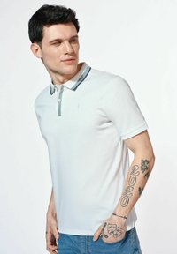 Biała koszulka polo z zapięciem na guziki, z kontrastowym jasno-niebieskim paskiem wzdłuż krawędzi kołnierza. Gładki materiał i krótkie rękawy.