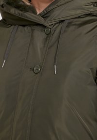 Olivově zelená bunda s kapucí lemovanou umělou kožešinou, druky, detaily na stažení a hladkým, lesklým povrchem.