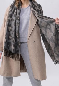 Personne portant un manteau beige et une tenue grise côtelée, tenant ouverte une grande écharpe imprimée animal gris et noir avec des bords à franges.