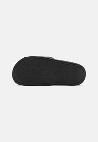 Suela de sandalia de goma negra con un patrón de agarre texturizado y el logo "LACOSTE" en relieve para una mayor visibilidad de la marca.