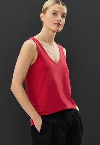 Top rosso senza maniche con scollatura a V, realizzato in tessuto morbido. La maglietta ha una vestibilità comoda ed è abbinata a pantaloni neri.