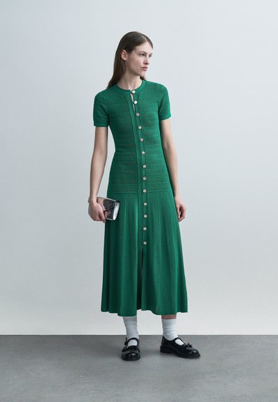 Femme portant une robe midi verte boutonnée, chaussettes blanches mi-mollet, mocassins noirs, tenant une petite pochette métallique, debout devant un fond gris uni.