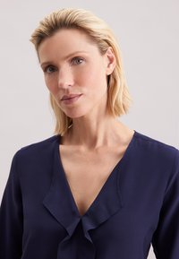 Vaaleahiuksinen nainen, jolla on lyhyet hiukset, pukeutuneena laivaston siniseen V-kaula-aukkoinen puseroon, jossa on röyhelöyksityiskohta. Hän katsoo hieman sivulle yksinkertaisen taustan edessä.