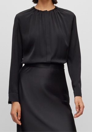 Femme portant un chemisier en satin noir à manches longues avec un col rond froncé et une jupe en satin noir assortie.
