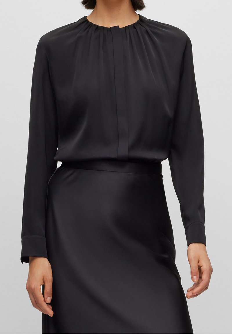 Femme portant un chemisier en satin noir à manches longues avec un col rond froncé et une jupe en satin noir assortie.
