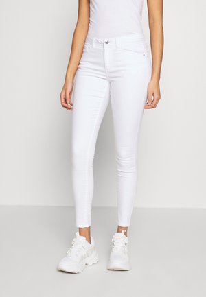Femme portant un jean blanc slim, des baskets blanches et un top blanc sans manches, debout devant un fond clair uni.