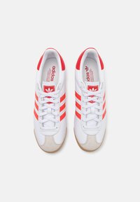 Λευκά αθλητικά παπούτσια με κόκκινες ρίγες, υφασμάτινη μύτη από υλικό σουέτ και σχέδιο με κορδόνια. Μάρκα ορατή στη γλώσσα και στην εσωτερική σόλα. Ελαστική σόλα.