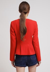 Femme avec une longue queue de cheval brune portant un blazer rouge ajusté et un short en denim bleu foncé, se tenant contre un fond clair uniforme.