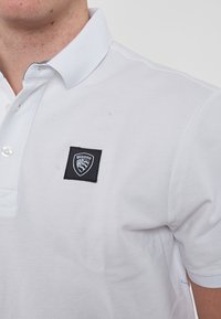 Polo bianco con tessuto testurizzato, dotato di una toppa quadrata nera con logo sul lato sinistro del petto e colletto cucito.