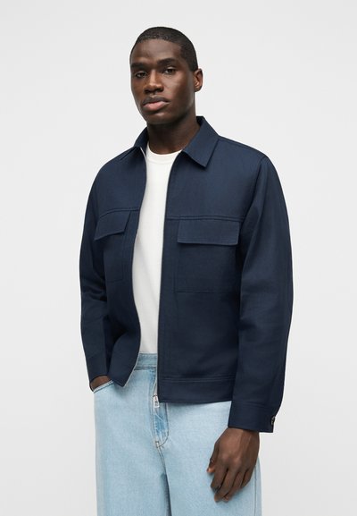 Άνδρας που φοράει ναυτικό μπλε μπουφάν με φερμουάρ πάνω από λευκό πουκάμισο και ανοιχτό μπλε τζιν, στέκεται μπροστά σε απλό ανοιχτό γκρι φόντο.