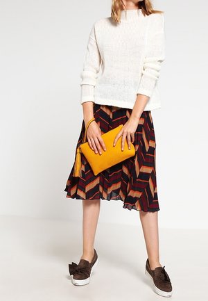 Jersey blanco de punto, falda estampada multicolor con chevrones rojos y marrones, bolso de mano amarillo con borla y zapatos marrones con lazos.