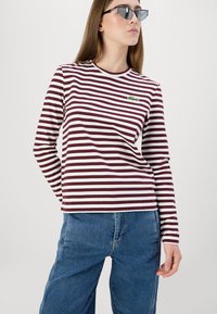 Camisa de manga larga a rayas en color rojo oscuro y blanco, con un pequeño logotipo verde en el pecho. Combinada con jeans de tiro alto en color azul.