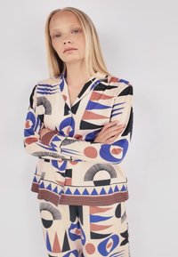 Pyjamaset met een beige stof met geometrische patronen in zwart, blauw, rood en crème; bevat een bovenstuk met kraag en bijpassende broek.