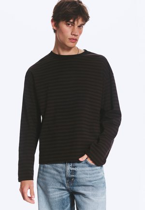 Young man with short brown hair wears dark long-sleeve striped shirt and light blue jeans, posing with one hand in pocket.
