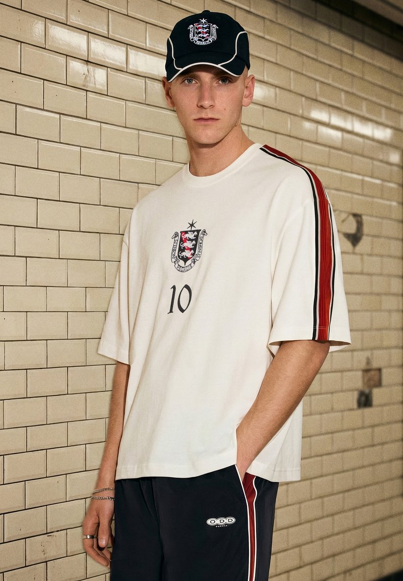 Jeune homme portant un maillot blanc avec le numéro 10 et un emblème, casquette noire avec un emblème assorti, debout contre un mur carrelé beige.