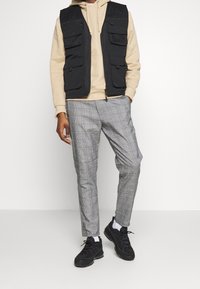 Black multi-pocket vest over a beige hoodie, paired with gray checkered trousers and black sneakers, featuring white socks and laces.