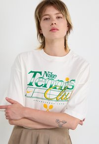 Valge puuvillane T-särk, millel on roheline ja kollane "Nike Tennis Club" graafika, lühikesed varrukad ja mugav lõige.
