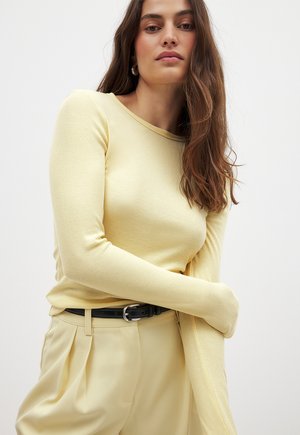 Gelbes geripptes Langarmshirt mit Rundhalsausschnitt, getragen mit hochtaillierten kremfarbigen Hosen und einem schwarzen Gürtel mit einer silbernen Schnalle.