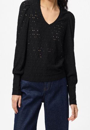 Kvinde iført en sort strikket trøje med lange ærmer med subtile blonde mønstre og blå højtaljede jeans mod en ensfarvet baggrund.
