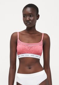 Roza bralette s čipkastim detaljima, prozirnom tkaninom, širokim belim pojasom s "GUESS" logotipom i tankim podesivim trakama. Glatka tekstura.