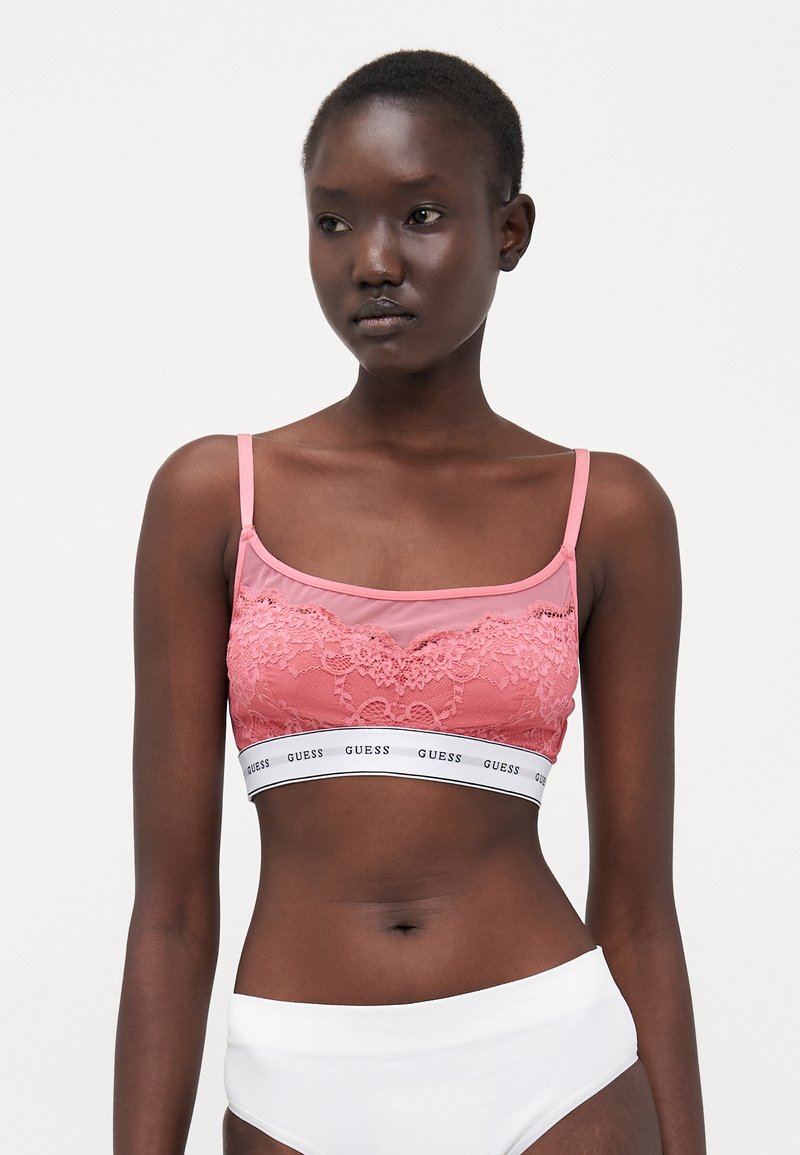 Roza bralette s čipkastim detaljima, prozirnom tkaninom, širokim belim pojasom s "GUESS" logotipom i tankim podesivim trakama. Glatka tekstura.