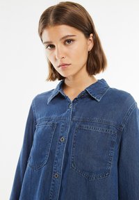 Denim srajca v temno modri barvi, z gumbnimi zapenjanjem spredaj, dvema žepoma na prsih in razpognjenim ovratnikom. Tkanina ima rahlo zbledel videz.