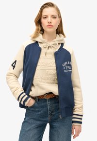 Chaqueta de cremallera en azul marino y crema con puños a rayas. Presenta una sudadera con capucha tipo canguro debajo, con texto en relieve y un cinturón tejido. Unos vaqueros de mezclilla completan el atuendo.