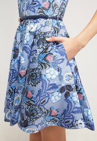 Blommig klänning med blå och svarta mönster på en ljusblå bakgrund. Inkluderar en sidoficka och en definierad midja med bälte.}}