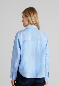 Camisa de botones de rayas azul claro con yugo en la parte posterior, mangas largas y un ajuste relajado. La tela parece lisa y ligera.