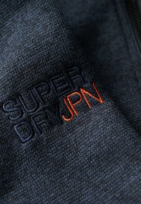 Un textile bleu marine foncé avec un motif tissé. "SUPERDRY" brodé en bleu marine et "JPN" en orange, positionné de manière bien en vue.