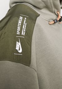 Sudadera color verde oliva con acento texturizado en los hombros, que presenta el logo blanco de Nike y la impresión "SPORTSWEAR". Cordón ajustable naranja en el cuello.