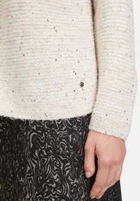 Hellbeige gestrickter Pullover mit Pailletten, der über eine strukturierte Oberfläche verfügt und am Saum mit einem silberfarbenen Detail versehen ist. Kombiniert mit einem gemusterten schwarzen Rock.