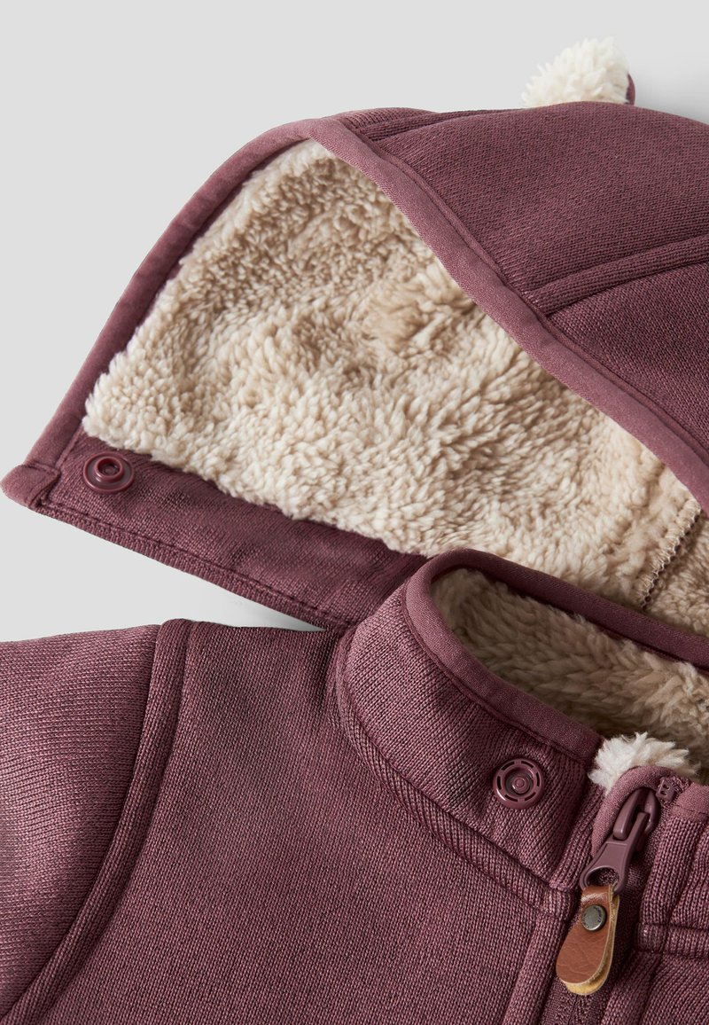 Bordeauxfarbene Kapuzenjacke mit plüschigem beigem Futter, ausgestattet mit einem Reißverschluss, Druckknopf und strukturiertem Außenmaterial. Pompon-Akzent an der Kapuze.