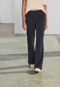 Juodos, plačios kelnės su lygia tekstūra, derinamos su smėlio spalvos palaidine ir šviesiais, atvirais avalynės pirštais. Paprasta diržo detalė ties juosmeniu.