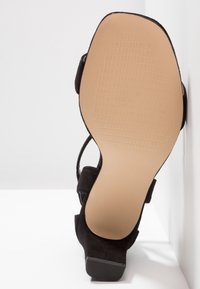 Sandale noire à bout ouvert avec une bride de cheville, vue par le dessous, présentant une semelle texturée beige et un petit talon carré.