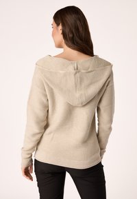 Hoodie in maglia di un beige chiaro con scollatura ampia e cappuccio con cordino. Presenta polsini e orlo a coste, evidenziando un tessuto testurizzato.