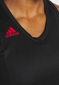 Camiseta sin mangas negra de deporte hecha de material liviano, con un escote en V profundo y el logo rojo de Adidas en el hombro izquierdo.