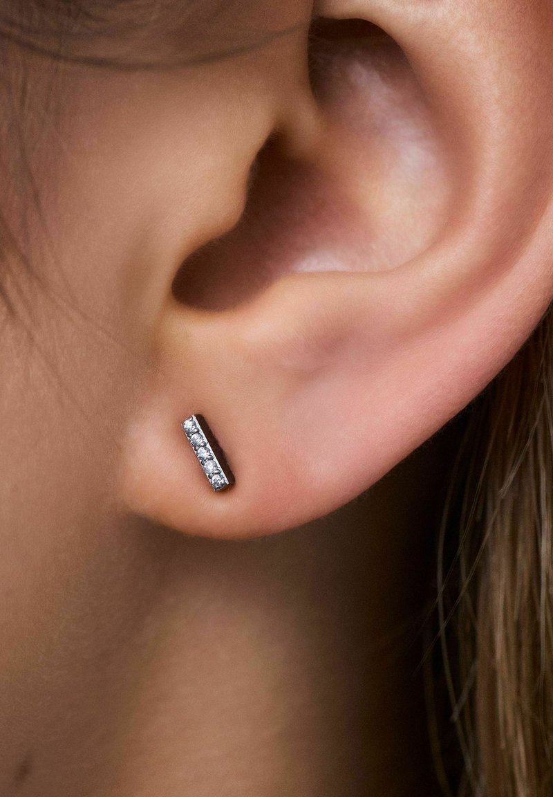 Clou d'oreille en argent de forme rectangulaire avec cinq petites pierres précieuses claires alignées en rangée, porté sur le lobe de l'oreille.