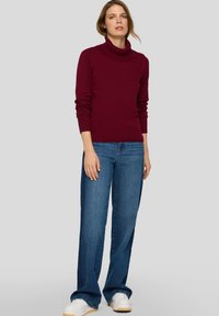 Weinroter Rollkragenpullover mit geripptem Saum und Knöpfen an den Ärmeln, kombiniert mit ausgestellten blauen Jeans und weißen Sneakern. Glatte Textur, lässige Passform.