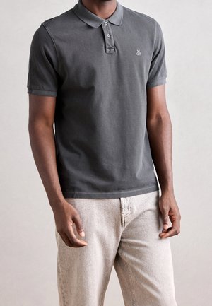 Man die een donkergrijs polo met korte mouwen en twee knopen draagt en lichtbeige broek, staand tegen een effen achtergrond.