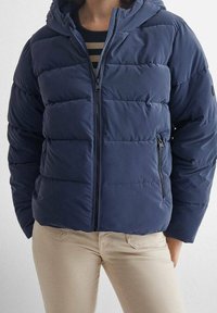 Osoba oblečená v tmavě modré prošívané bundě s kapucí přes pruhované tričko a béžové kalhoty, stojící s rukama částečně v kapsách.