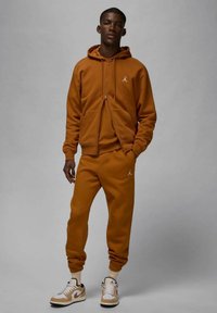 Felpa chiusa con zip marrone e pantaloni abbinati con polsini a costine. Logo bianco sul lato sinistro del petto e sui pantaloni. Sneakers con dettagli beige e bianchi.