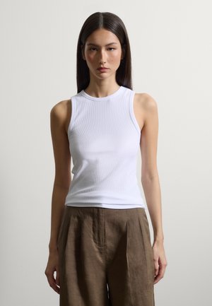 Jeune femme aux longs cheveux foncés portant un haut blanc sans manches et un pantalon marron ample, debout devant un fond clair uni.