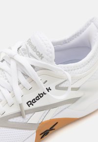 Bílé atletické boty Reebok s síťovinou, bílými tkaničkami, stříbrnými detaily a béžovou podrážkou, na které je logo Reebok.