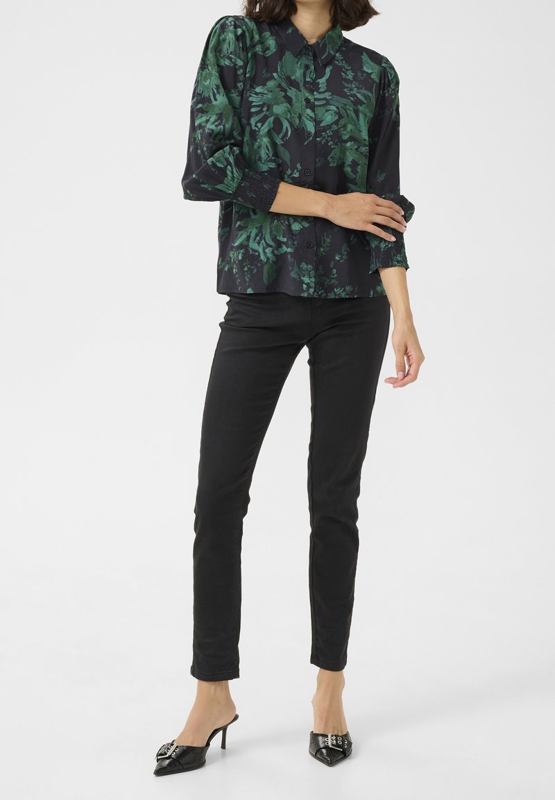 Femme portant un pantalon noir, une chemise à manches longues fleurie vert foncé et des chaussures à talons hauts pointues noires, debout sur un fond blanc.