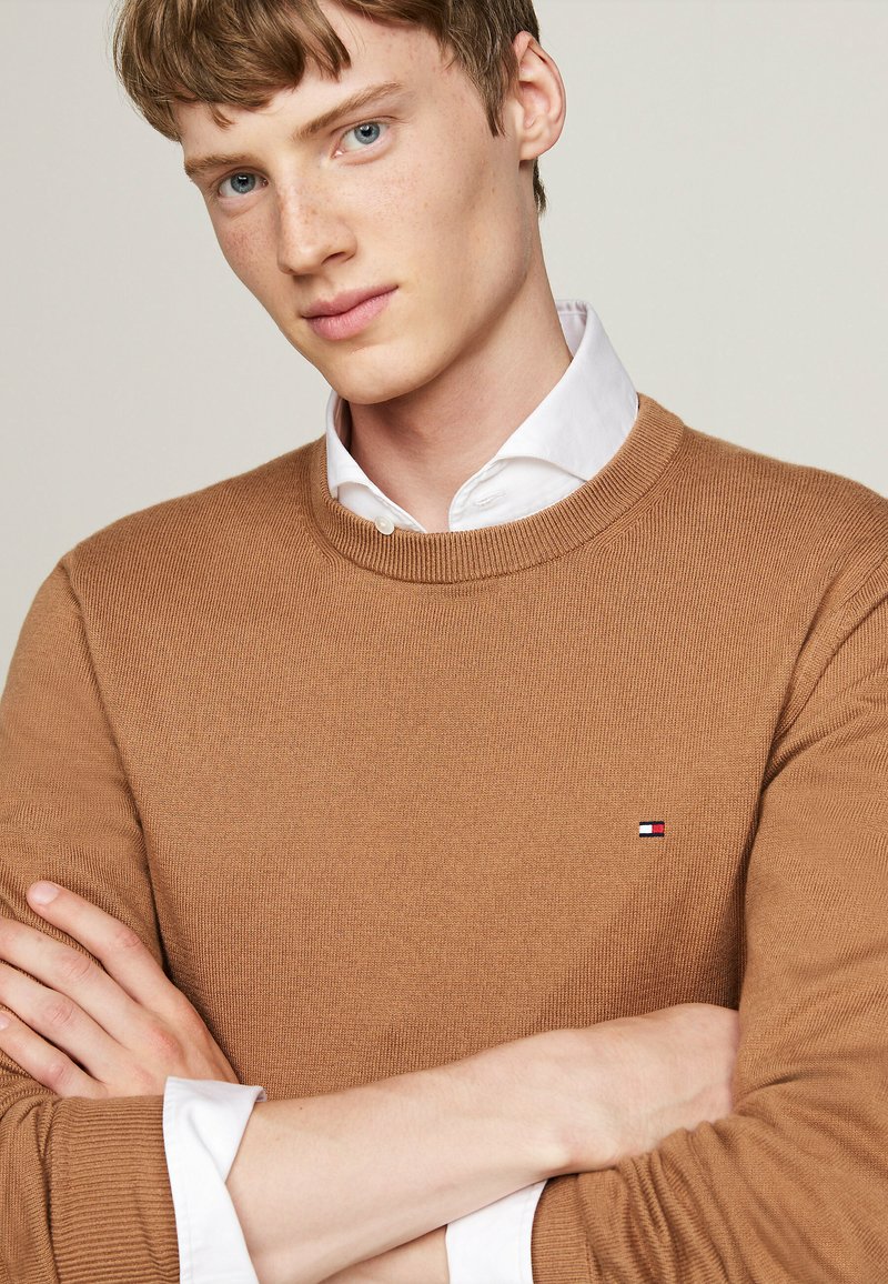 Junger Mann mit heller Haut und Sommersprossen, der ein weißes Hemd unter einem braunen Pullover trägt, mit verschränkten Armen und leicht zur Seite blickend.