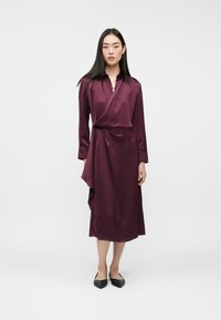 Burgundowa satynowa sukienka w stylu wrap z długimi rękawami, kołnierzykiem i drapowaniem w talii, połączona z czarnymi czółenkami w szpic.