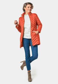 Gequilte oranje jas met hoge kraag en gouden ritsdetail, gecombineerd met een witte top en blauwe jeans, tegen een witte achtergrond.