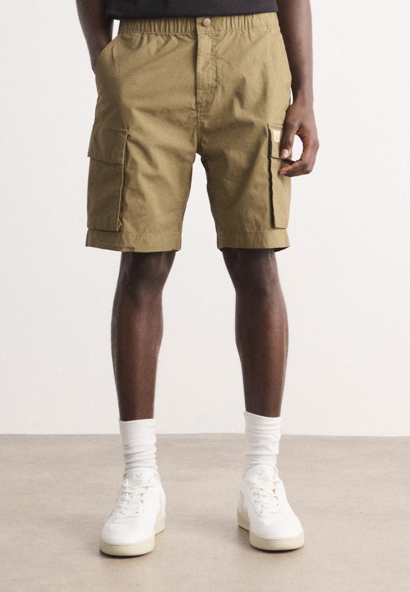 Pantaloni cargo color khaki con cintura elastica, due tasche laterali e taglio a ginocchio. Abbinati a sneakers alte bianche e calze.