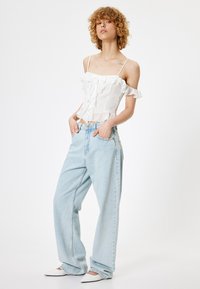 Weißes schulterfreies Oberteil mit fransigen Kanten, kombiniert mit hellblauen, weiten Jeans und spitzen weißen Schuhen. Schlichtes, sauberes Design.